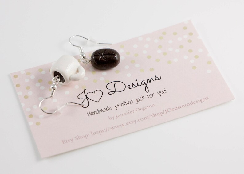 May include: A pair of earrings featuring a white ceramic coffee cup and a brown chocolate-colored donut. The earrings are on a pink background with gold polka dots. The text "J Designs" is written in black cursive script. The text "Handmade pretties just for you!" is written in black. The text "by Jennifer Orgeron" is written in black. The text "Etsy Shop: https://www.etsy.com/shop/JOcustomdesigns" is written in black.