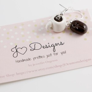 May include: A pair of handmade earrings featuring a white ceramic coffee cup and a brown coffee bean. The earrings are on a pink background with white polka dots. The text "J Designs Handmade pretties just for you! by Jennifer Orgeron Etsy Shop: https://www.etsy.com/shop/JOcustomdesigns" is printed on the background.