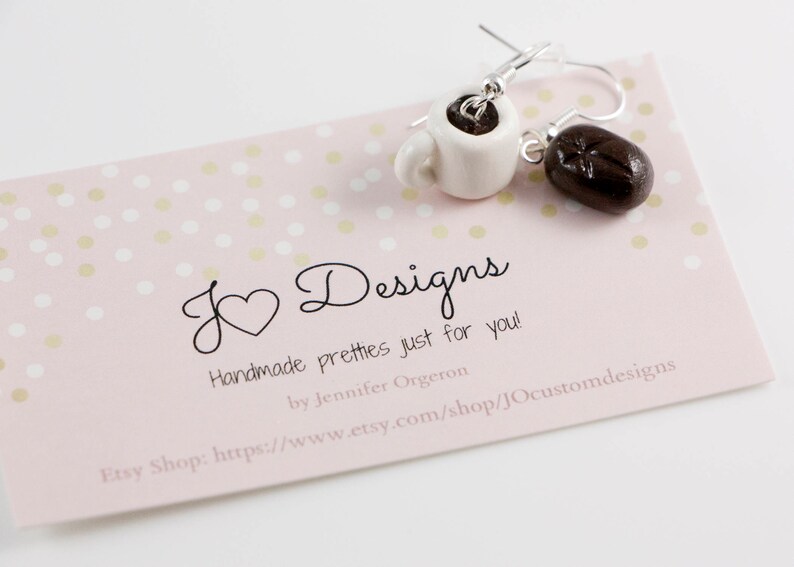 May include: A pair of handmade earrings featuring a white ceramic coffee cup and a brown coffee bean. The earrings are on a pink background with white polka dots. The text "J Designs Handmade pretties just for you! by Jennifer Orgeron Etsy Shop: https://www.etsy.com/shop/JOcustomdesigns" is printed on the background.
