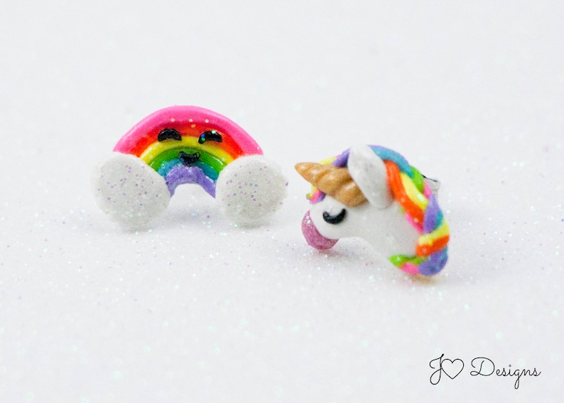 May include: A pair of colorful rainbow and unicorn earrings. The rainbow earring is pink with a smiling face and white clouds. The unicorn earring is white with a rainbow mane and horn.
