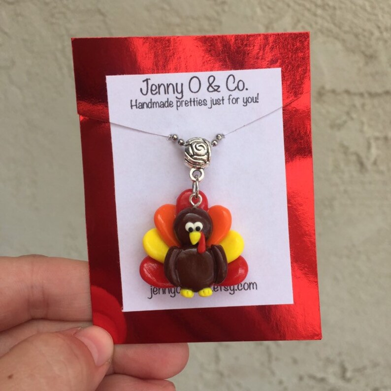 May include: A handmade polymer clay turkey charm necklace on a silver chain with a silver bead. The charm is brown with a red, orange, and yellow feathered tail. The necklace is displayed on a red card with the text "Jenny O & Co. Handmade pretties just for you!" and "jennyo.etsy.com".