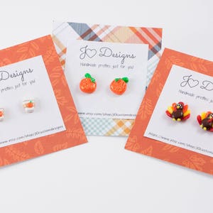 May include: Three cards with fall-themed earrings. The cards are orange with a white border and feature the text "J Designs Handmade pretties just for you". The earrings are a pair of orange pumpkins, a pair of orange cups with lids, and a pair of turkeys.