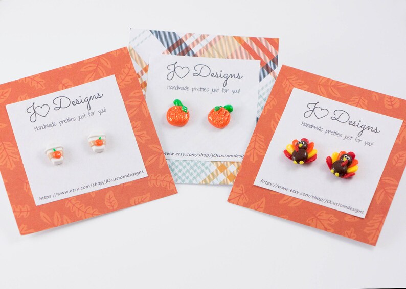 May include: Three cards with fall-themed earrings. The cards are orange with a white border and feature the text "J Designs Handmade pretties just for you". The earrings are a pair of orange pumpkins, a pair of orange cups with lids, and a pair of turkeys.