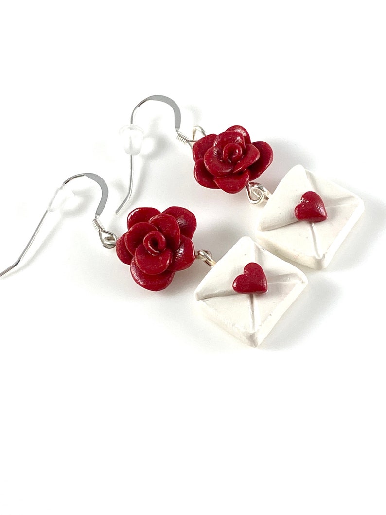 May include: A pair of dangle earrings featuring a red rose charm and a white envelope charm with a red heart on the front.