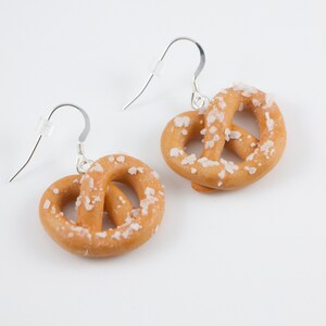 Puede incluir: Un par de pendientes de aro de plata con un dije en forma de pretzel. El pretzel es marr&oacute;n y tiene una capa de sal blanca.