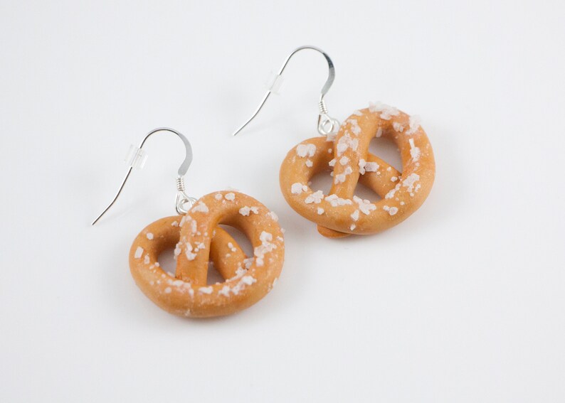 Puede incluir: Un par de pendientes de aro de plata con un dije en forma de pretzel. El pretzel es marr&oacute;n y tiene una capa de sal blanca.