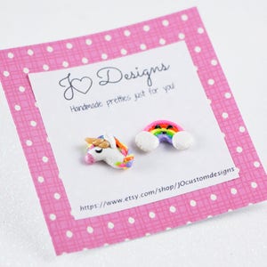 May include: A pink card with white polka dots and the text "J&hearts;Designs Handmade pretties just for you" and a website address. The card has two small clay charms attached to it: a rainbow with clouds and a unicorn.