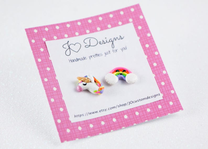 May include: A pink card with white polka dots and the text "J&hearts;Designs Handmade pretties just for you" and a website address. The card has two small clay charms attached to it: a rainbow with clouds and a unicorn.