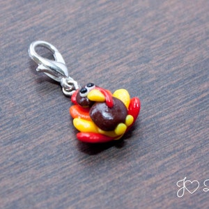 May include: A small, brown turkey charm with red and yellow feathers. The charm is attached to a silver metal clasp.