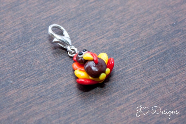 May include: A small, brown turkey charm with red and yellow feathers. The charm is attached to a silver metal clasp.