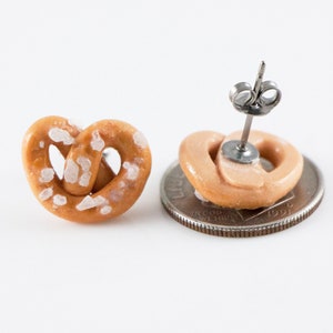 May include: A pair of salt pretzel earrings. The earrings are made of polymer clay and are shaped like pretzels. The pretzels are brown and have white salt crystals on them. The earrings are attached to silver posts.