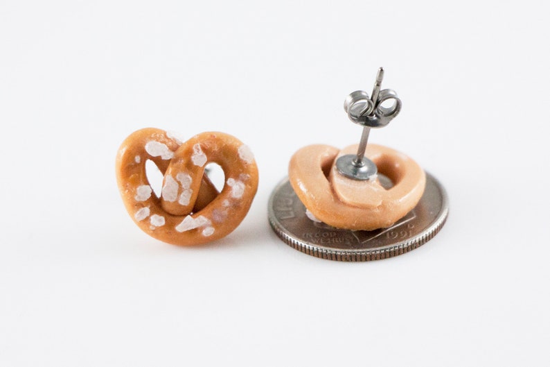 May include: A pair of salt pretzel earrings. The earrings are made of polymer clay and are shaped like pretzels. The pretzels are brown and have white salt crystals on them. The earrings are attached to silver posts.