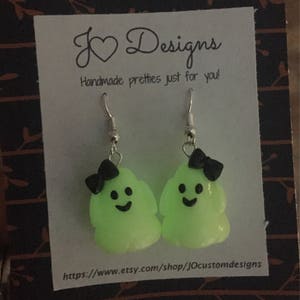 May include: A pair of green glow-in-the-dark ghost earrings with black bows. The earrings are on a white card with the text "J &hearts; Designs Handmade pretties just for you!" and a website address.