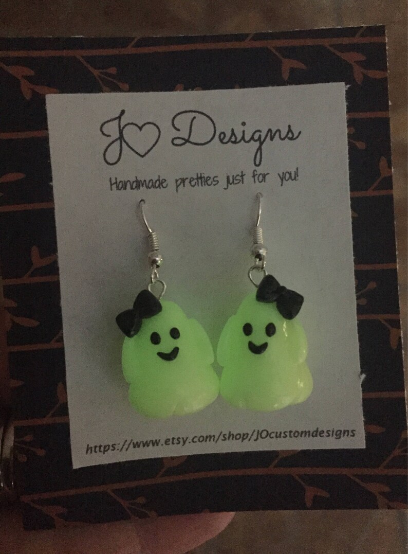 May include: A pair of green glow-in-the-dark ghost earrings with black bows. The earrings are on a white card with the text "J &hearts; Designs Handmade pretties just for you!" and a website address.