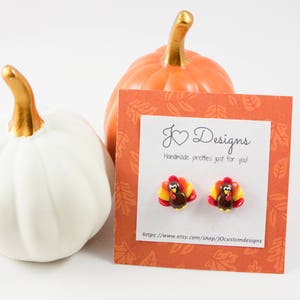 May include: A pair of polymer clay earrings featuring a cute turkey design. The earrings are brown, red, yellow, and orange. The earrings are displayed on a card with the text "J &hearts; Designs Handmade pretties just for you" and a website address.