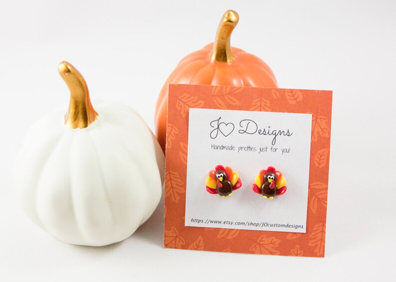 May include: A pair of polymer clay earrings featuring a cute turkey design. The earrings are brown, red, yellow, and orange. The earrings are displayed on a card with the text "J &hearts; Designs Handmade pretties just for you" and a website address.