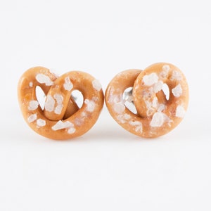 May include: A pair of salt-covered pretzel earrings. The earrings are made of polymer clay and are a light brown color.