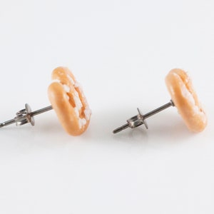 May include: A pair of gold-colored earrings shaped like a donut with white frosting. The earrings are made of polymer clay and have a silver post.