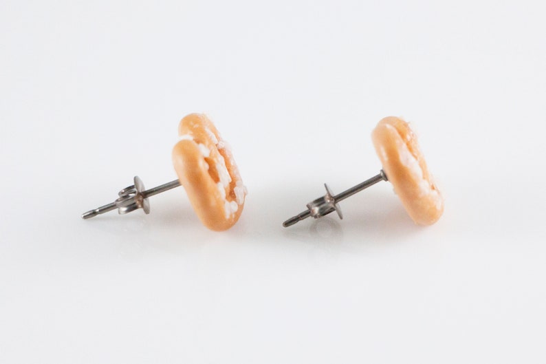 May include: A pair of gold-colored earrings shaped like a donut with white frosting. The earrings are made of polymer clay and have a silver post.