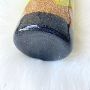 Puede incluir: Un vaso de glitter negro y dorado con el texto "Get Erased" en la parte superior. El vaso est&aacute; sobre una superficie blanca y esponjosa.