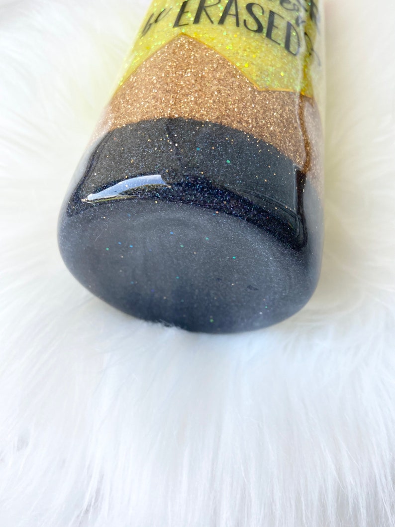 Puede incluir: Un vaso de glitter negro y dorado con el texto "Get Erased" en la parte superior. El vaso est&aacute; sobre una superficie blanca y esponjosa.