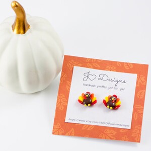 May include: A pair of small, handmade turkey earrings. The earrings are brown, red, and orange with a gold heart charm. The earrings are displayed on a card with the text "J Designs Handmade pretties just for you" and a website address.