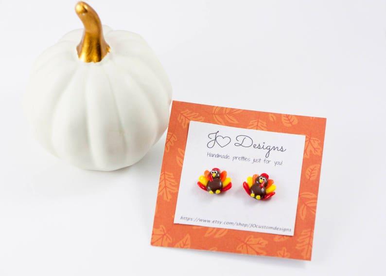 May include: A pair of small, handmade turkey earrings. The earrings are brown, red, and orange with a gold heart charm. The earrings are displayed on a card with the text "J Designs Handmade pretties just for you" and a website address.