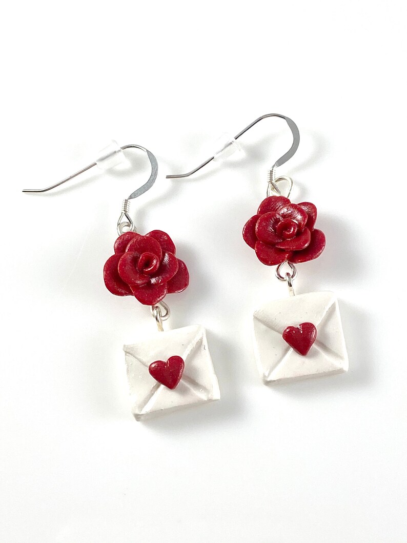 May include: A pair of dangle earrings featuring a red rose and a white envelope with a red heart on each earring.