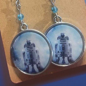 May include: A pair of silver dangle earrings with a blue glass bead on each earring. The earrings feature a round, silver-rimmed charm with a printed image of R2-D2 from Star Wars.