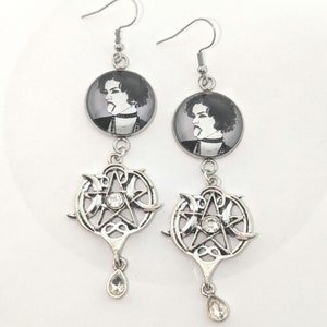 May include: Silver dangle earrings with a black and white image of a person's face on the top and a silver pentagram with a crystal in the center on the bottom.