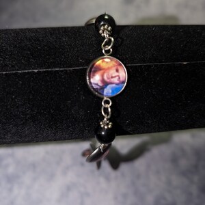 May include: A silver bracelet with black beads and a round charm featuring a colourful image of a person's face.