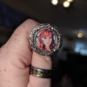 May include: A silver ring with a cameo featuring a woman with red hair. The cameo is surrounded by an ornate silver design.
