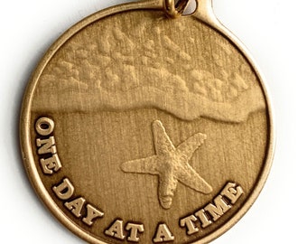 May include: Gold-toned keychain with a round charm featuring a starfish and the text "ONE DAY AT A TIME".