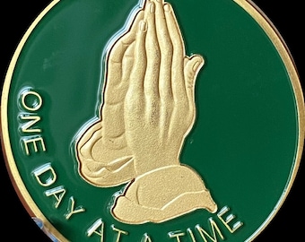 May include: A green round coin with a gold image of two hands clasped in prayer. The text "ONE DAY AT A TIME" is written around the image.