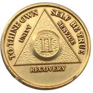 Puede incluir: Moneda de metal dorado con un diseño triangular y las palabras "To Thine Own Self Be True" y "Unity" en la parte superior izquierda, "Service" en la parte superior derecha y "Recovery" en la parte inferior. El número "II" está en el centro del triángulo.