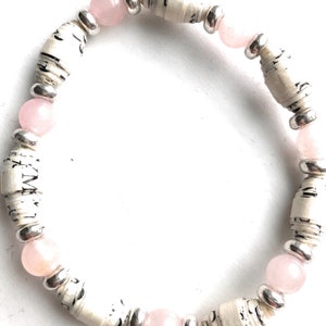 May include: A bracelet made of rolled paper beads and pink gemstone beads. The paper beads are white with black text and the gemstone beads are a light pink color.