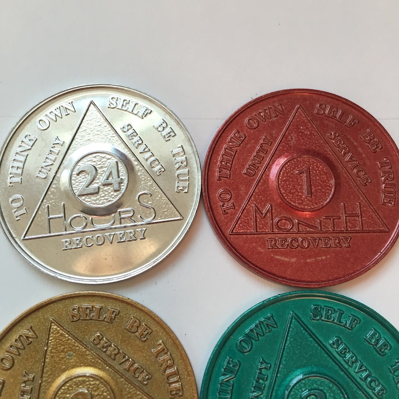 May include: Four sobriety coins with a triangle design. The coins are silver, gold, red, and green. Each coin has the text "To thine own self be true, Unity, Service" and a number representing the length of sobriety. The coins read "24 Hours", "1 Month", "2 Weeks", and "1 Year".