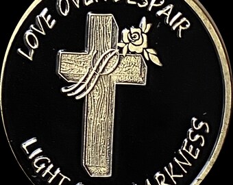 Puede incluir: Una moneda de color dorado con fondo negro. La moneda presenta una cruz de madera con una rosa y una cinta. El texto "LOVE OVER DESPAIR LIGHT OVER DARKNESS" está inscrito alrededor del borde de la moneda.