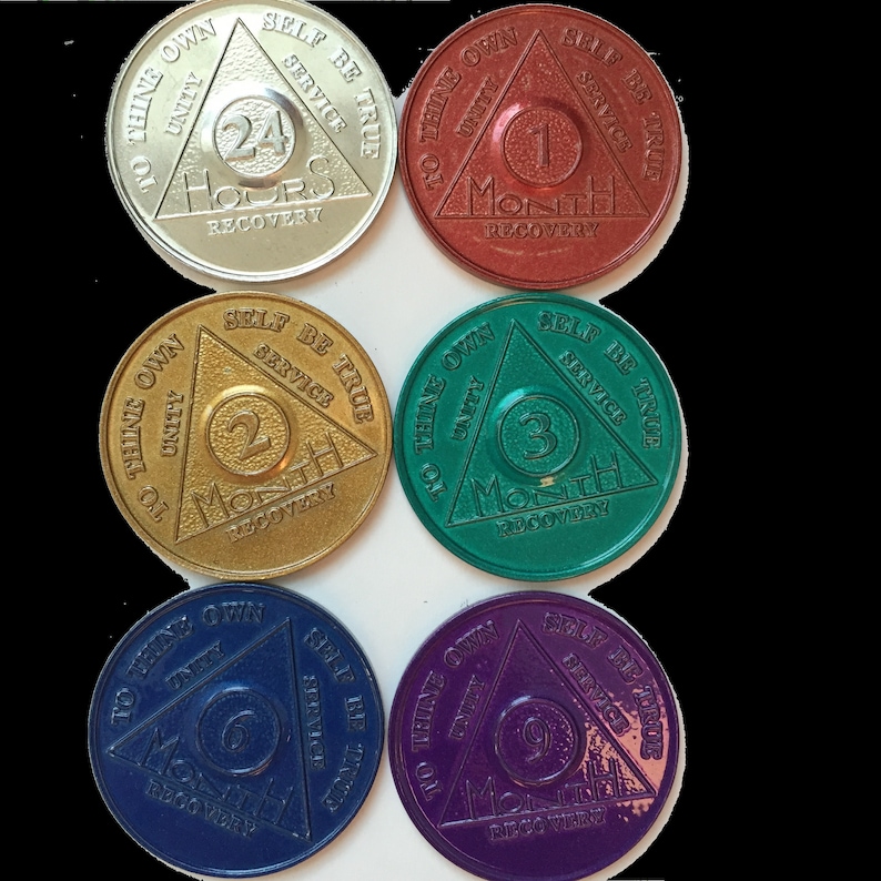 May include: Six round, metallic tokens with a triangle design. Each token features a different color and number, representing a time period of recovery: 24 hours, 1 month, 2 months, 3 months, 6 months, and 9 months. The text on each token reads "To thine own self be true, Unity, Service".