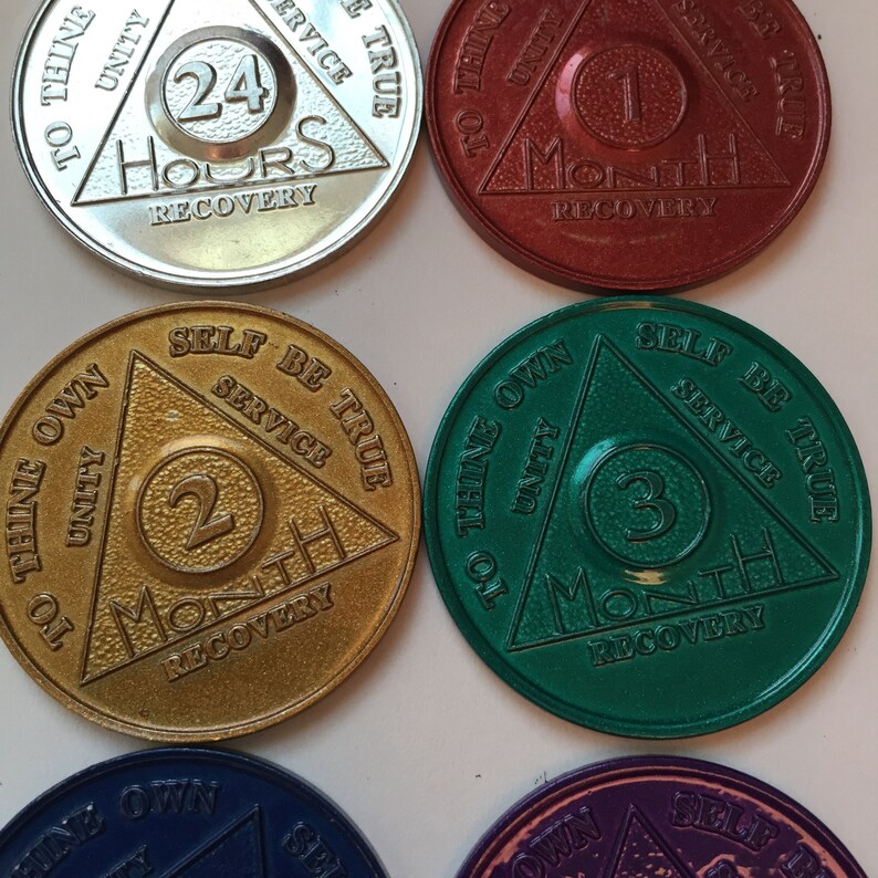 May include: Six sobriety coins with a triangular design. The coins are in different colors, including silver, red, gold, green, and purple. Each coin has a number in the center, representing the number of months of sobriety. The text on the coins includes "To Thine Own", "Unity", "Self Be True", "Service", and "Recovery".