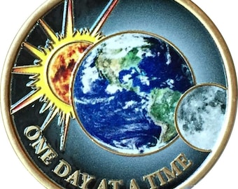 May include: A gold-coloured coin featuring a blue and green image of Earth, a yellow sun, and a grey moon. The text "ONE DAY AT A TIME" is inscribed around the edge of the coin.