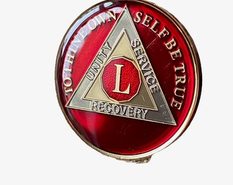 May include: A red, gold, and silver Alcoholics Anonymous (AA) sobriety coin with the words "To Thine Own Self Be True" and a triangle with the words "Unity, Service, Recovery" and the letter "L" in the center.