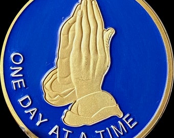 Puede incluir: Una moneda de color dorado con fondo azul. La moneda presenta un par de manos juntas en oración y el texto "ONE DAY AT A TIME".