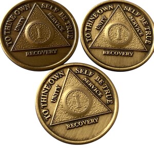 Può includere: Tre monete di metallo color oro con un design triangolare e le parole "To Thine Own", "Unity", "Self Be True", "Service" e "Recovery" incise su di esse.