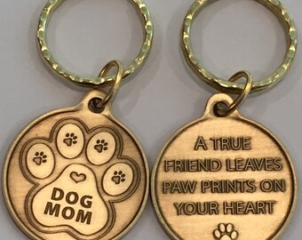 May include: Two bronze dog tag keyrings. One tag has a paw print design with the words "DOG MOM." The other tag has the inscription "A TRUE FRIEND LEAVES PAW PRINTS ON YOUR HEART" and a paw print.