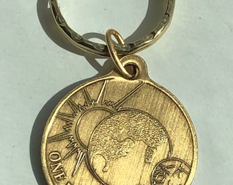 May include: A gold-toned keychain with a round charm featuring a sun, earth, and clock design. The charm has the text "ONE DAY AT A TIME" inscribed around the edge.
