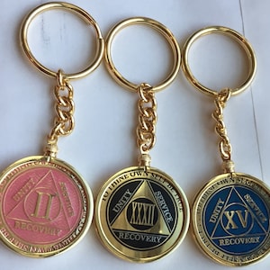 Könnte beinhalten: Drei goldene Schlüsselanhänger mit runden Medaillons, die ein Dreiecksdesign mit den Worten "To Thine Own Self Be True" und den Zahlen "II", "XXXII" und "XV" im Inneren zeigen. Die Medaillons sind rosa, schwarz und blau.