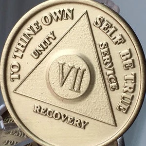 May include: Gold-colored metal coin with a triangle design and the words "To Thine Own Self Be True" and "Unity" inside the triangle. The words "Recovery" and "Service" are below the triangle. The number "VII" is in the center of the coin.
