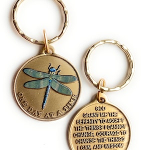 May include: Two gold-tone keychains. One features a dragonfly with blue and green wings and the text "One Day At A Time". The other keychain has the text "God Grant Me The Serenity To Accept The Things I Cannot Change, Courage To Change The Things I Can, And Wisdom To Know The Difference."
