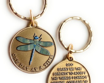 May include: Two gold-tone keyrings. One features a dragonfly with blue and green wings and the text "One Day At A Time". The other keyring has the text "God Grant Me The Serenity To Accept The Things I Cannot Change, Courage To Change The Things I Can, And Wisdom To Know The Difference."